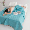 A teal breathable cooling blanket is draped over a white bed, with a person lying on it and an open booklet nearby.