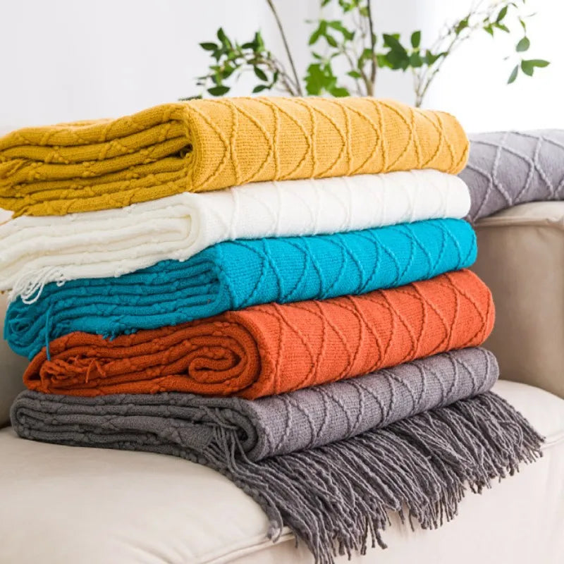 A stack of colorful knitted tassel throw blankets in yellow, cream, blue, teal, orange, and gray.