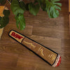 A rectangular, cigarette-shaped floor mat with a raw-edge design rests on a wooden floor surrounded by green leaves.