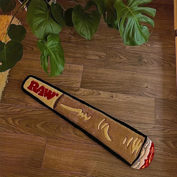 A rectangular, cigarette-shaped floor mat with a raw-edge design rests on a wooden floor surrounded by green leaves.