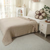 A beige quilted blanket is draped over a white bed, next to a white nightstand with a floral decorative item.