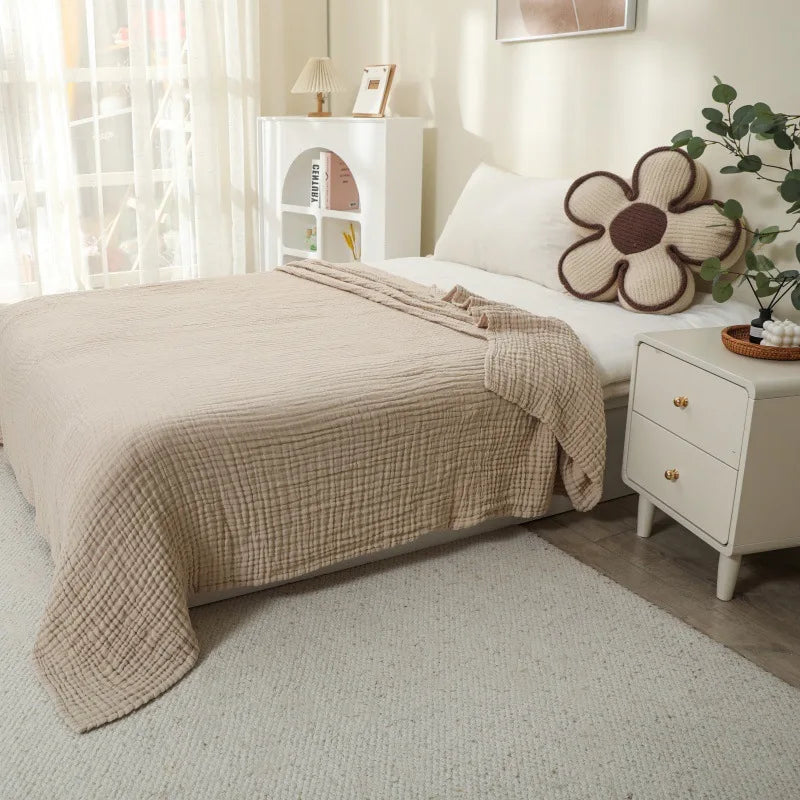 A beige quilted blanket is draped over a white bed, next to a white nightstand with a floral decorative item.