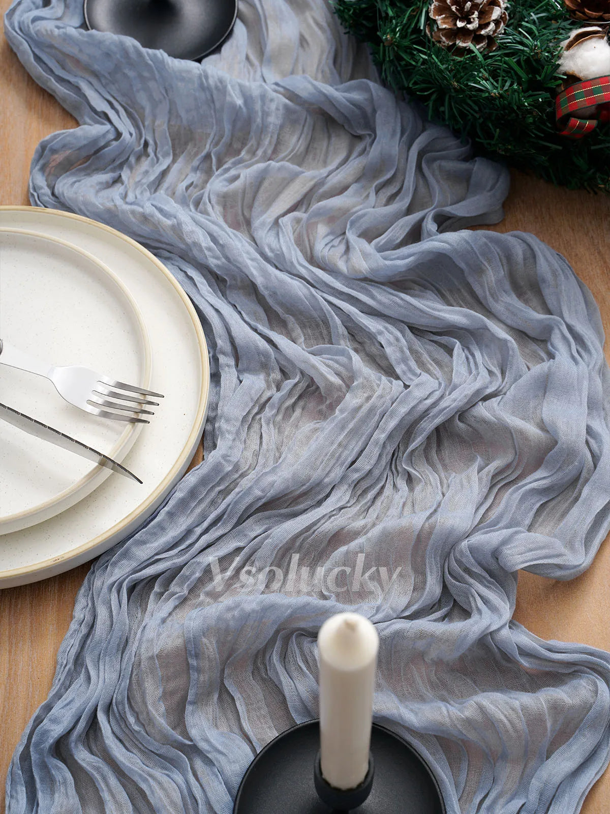 A gray crinkled cheesecloth table runner draped over a wooden surface with plates and a pine wreath.
