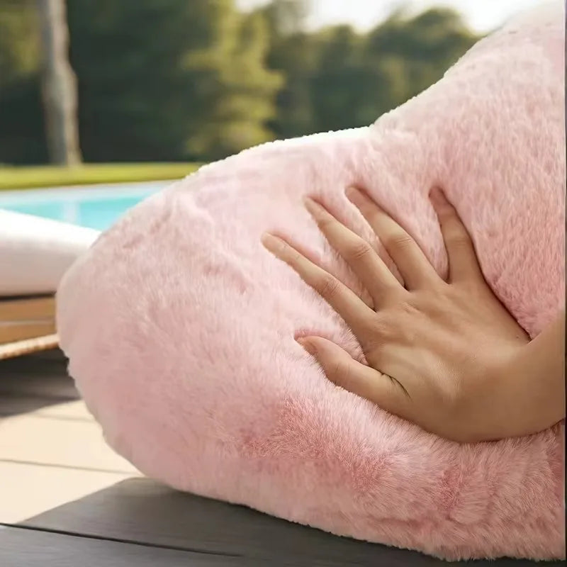 A pink heart-shaped plush cushion resting on a wooden surface with a hand touching it.