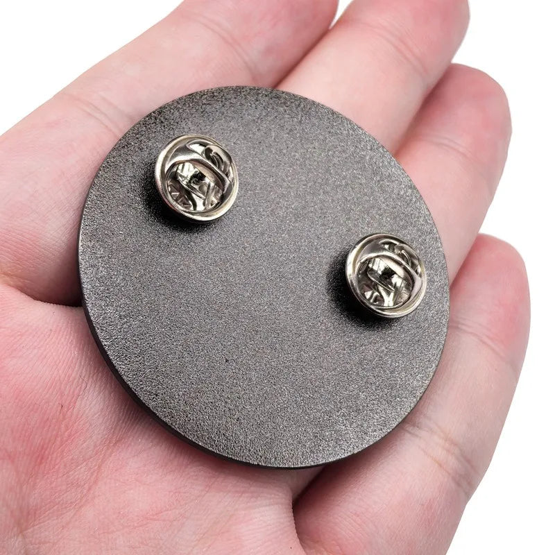 A dark gray metal brooch pin featuring Ho-Oh and Lugia designs is held in a person's hand.