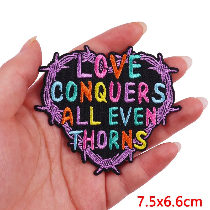 A hand holds a black embroidered patch with purple thorns and colorful text that reads Love Conquers All Even Thorns