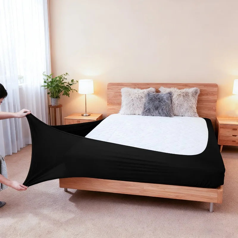 Black high-stretch elastic box spring cover placed on a wooden bed frame with fluffy pillows.