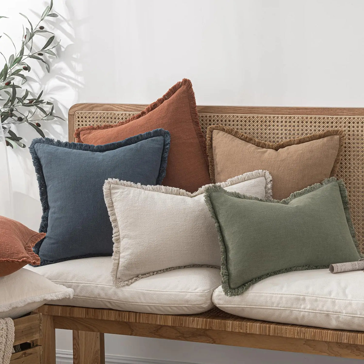 A collection of linen fringe throw pillow covers in earthy tones like terracotta, olive green, and beige resting on a wooden 