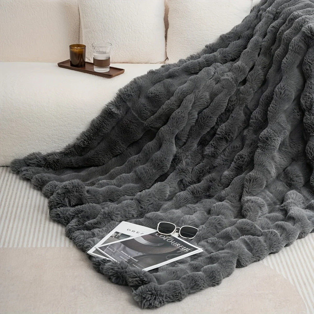 A dark gray luxury faux rabbit fur throw blanket draped over a white sofa with a glass and a wooden tray nearby.