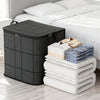 A black portable folding storage bag is placed next to a white bed with folded white and blue textiles.