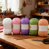 A collection of soft bulky chenille yarn in white, pink, purple, and green is displayed on a wooden table.