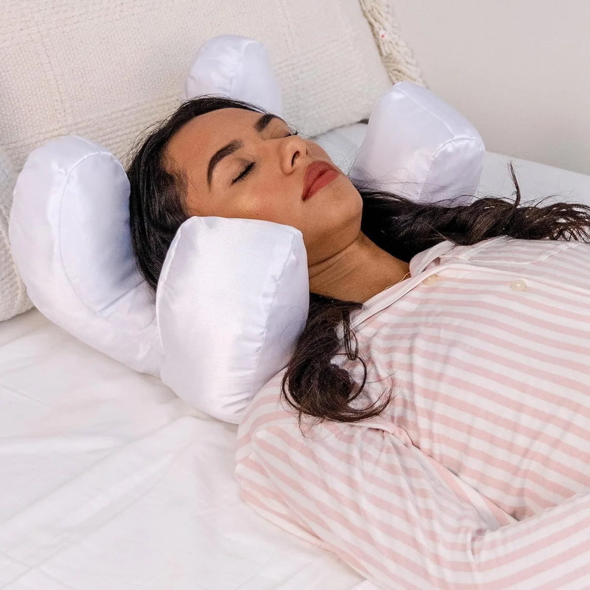 A woman is resting her head on a white pillow surrounded by several white satin anti-wrinkle pillows.
