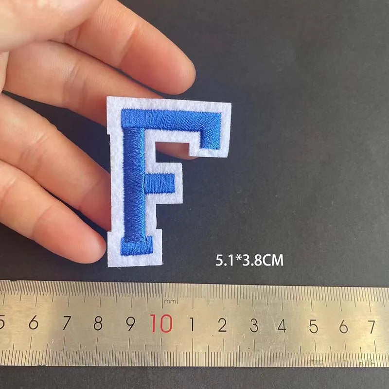 A hand holds a blue alphabet embroidery iron-on patch shaped like the letter F, measured against a ruler.