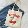 A beige canvas shopping bag featuring a red cherry design and the text Happy Have a Nice Day, placed on a white sheet next to