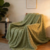 A textured, olive green plush throw blanket draped on a surface next to a potted plant.