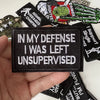 A hand holds a black iron-on slogan patch with white text that reads In My Defense I Was Left Unsupervised.