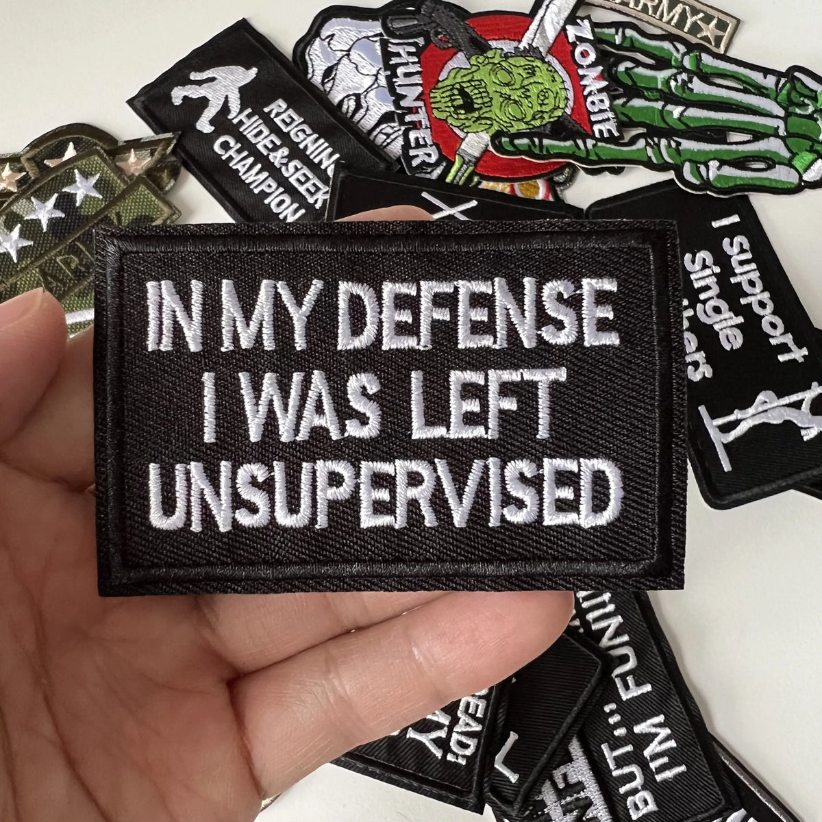 A hand holds a black iron-on slogan patch with white text that reads In My Defense I Was Left Unsupervised.