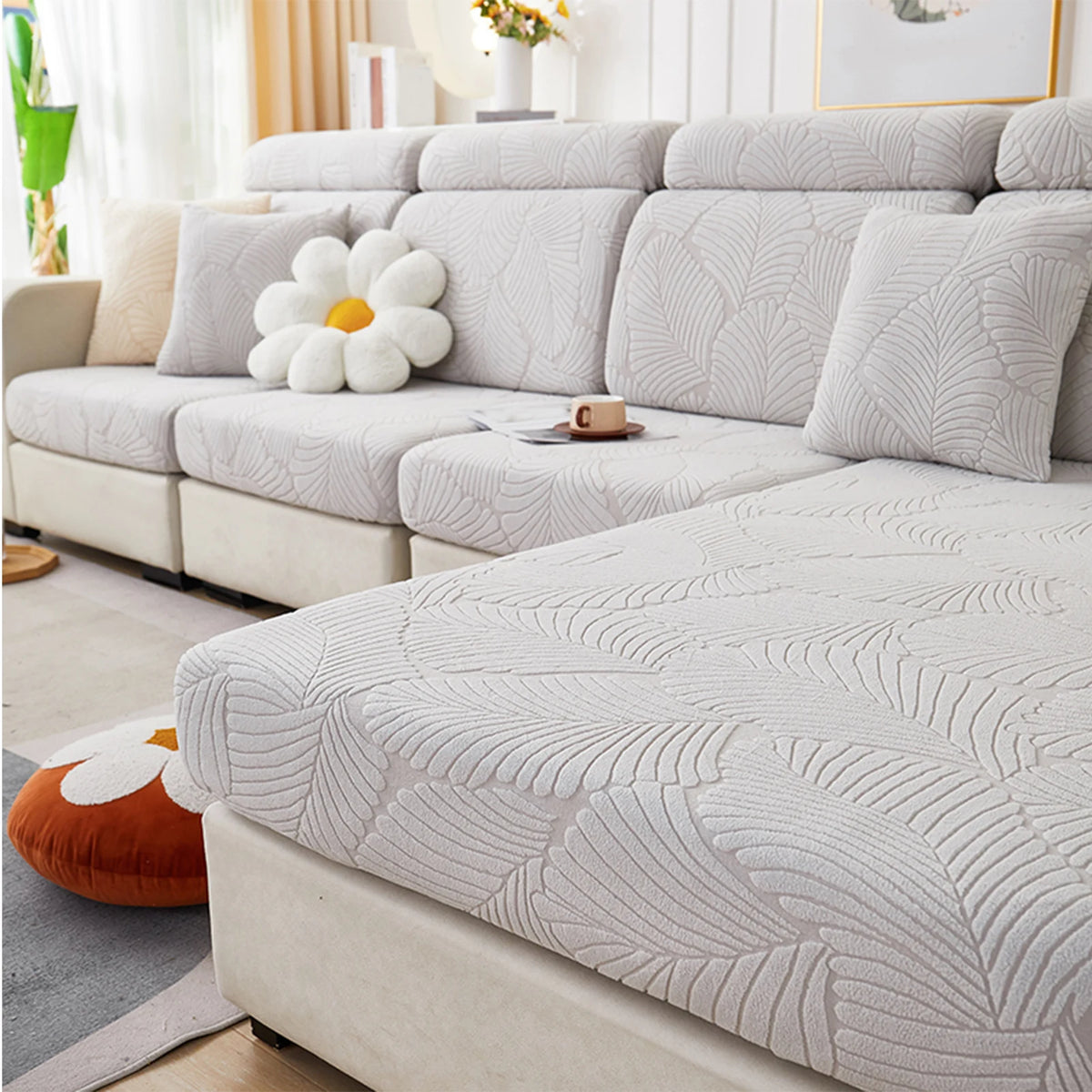 A light gray leaf jacquard stretch sofa cover displayed on a modern sectional sofa with white pillows.