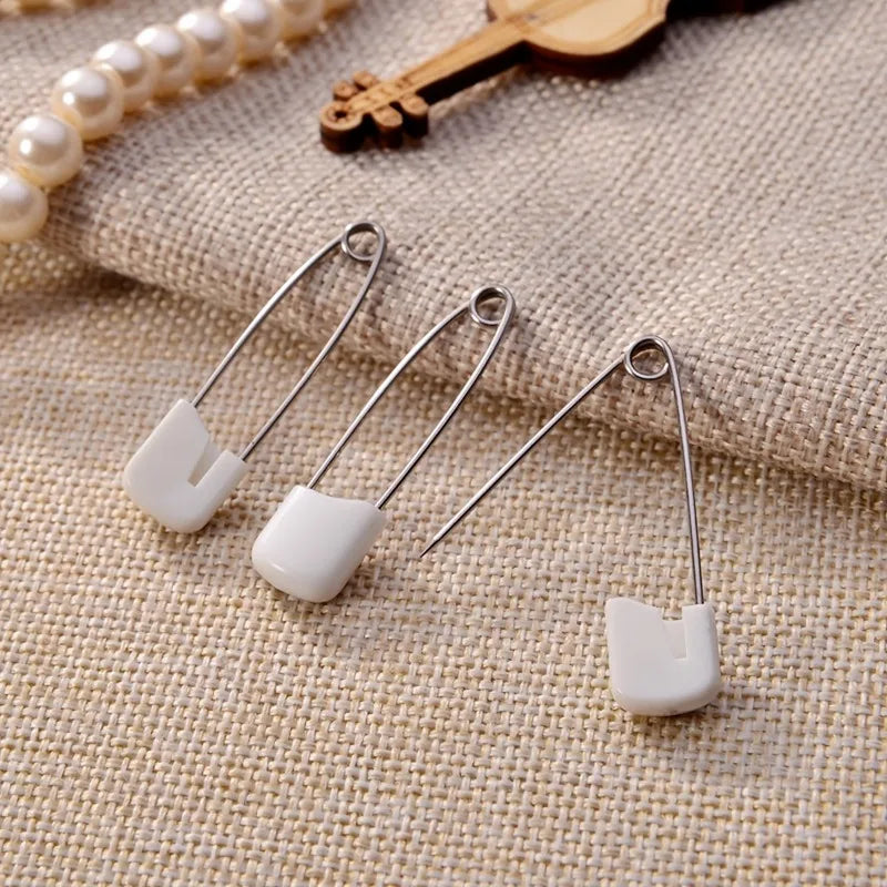 A close-up of stainless steel diaper pins with safety locking closures resting on a beige fabric.
