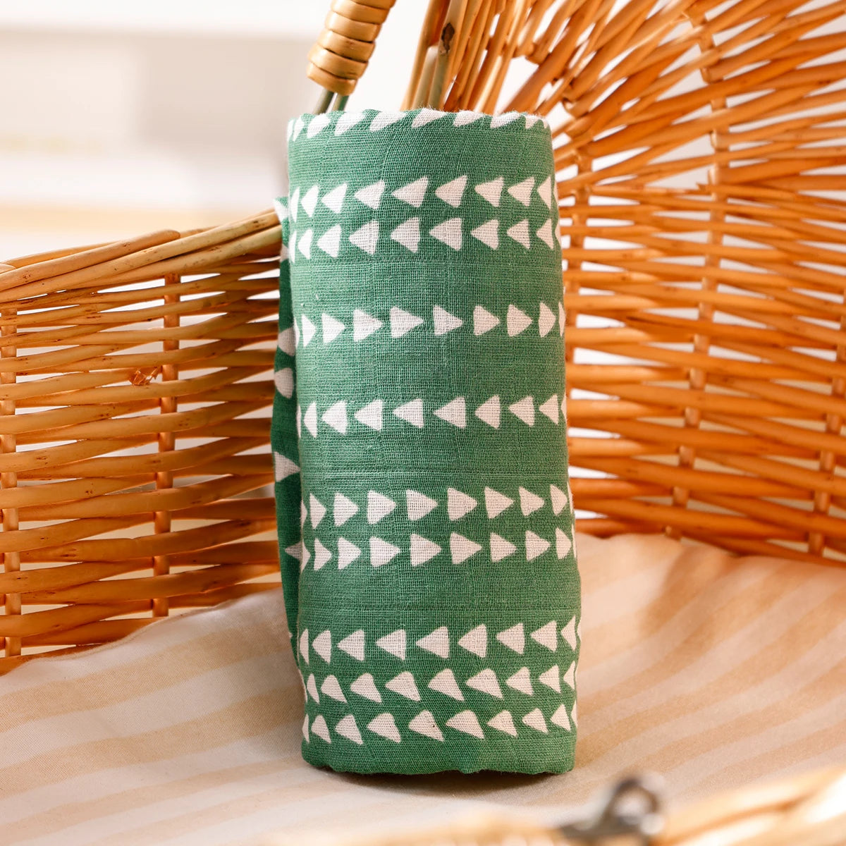 A green muslin swaddle blanket with white triangles is placed next to a woven basket.