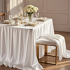 A white satin table overlay cloth is draped on a table set with elegant floral arrangements and glassware.