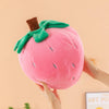 A pink plush strawberry throw pillow with green leaves is being held by a hand.