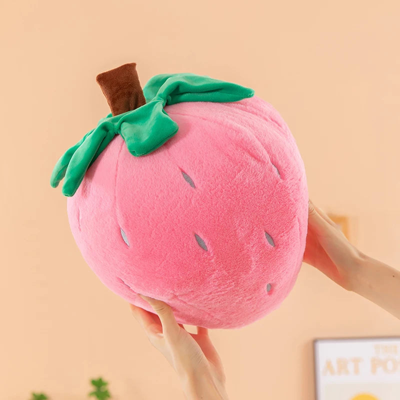 A pink plush strawberry throw pillow with green leaves is being held by a hand.