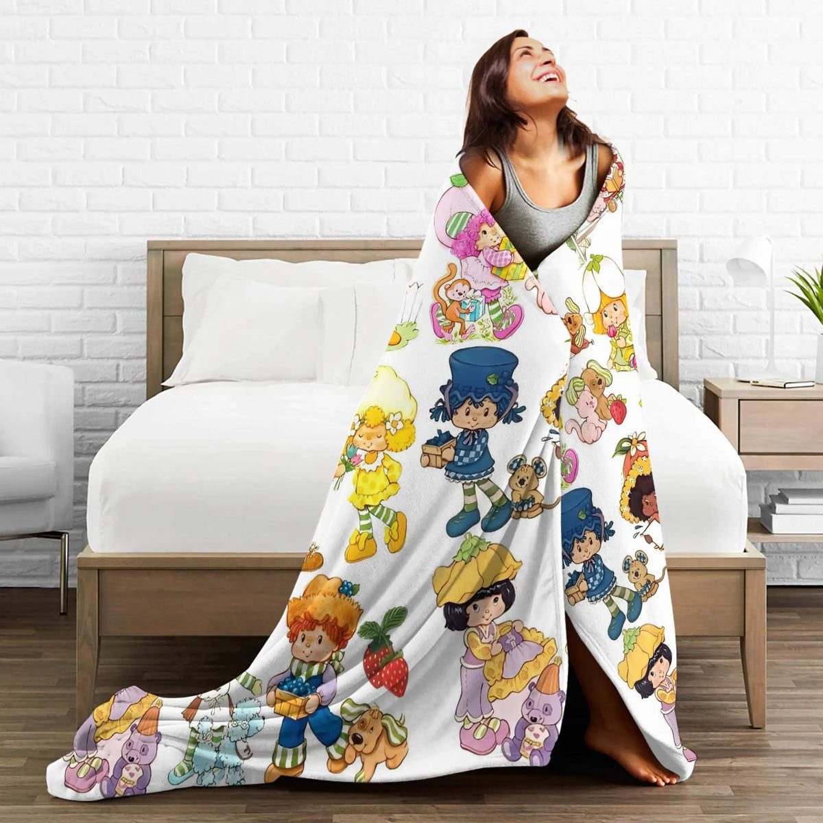A woman is smiling while wrapped in a large, colorful plush throw blanket featuring Strawberry Shortcake characters.