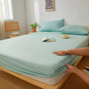A light mint green fitted sheet is laid out on a wooden bed frame with a hand touching the fabric.