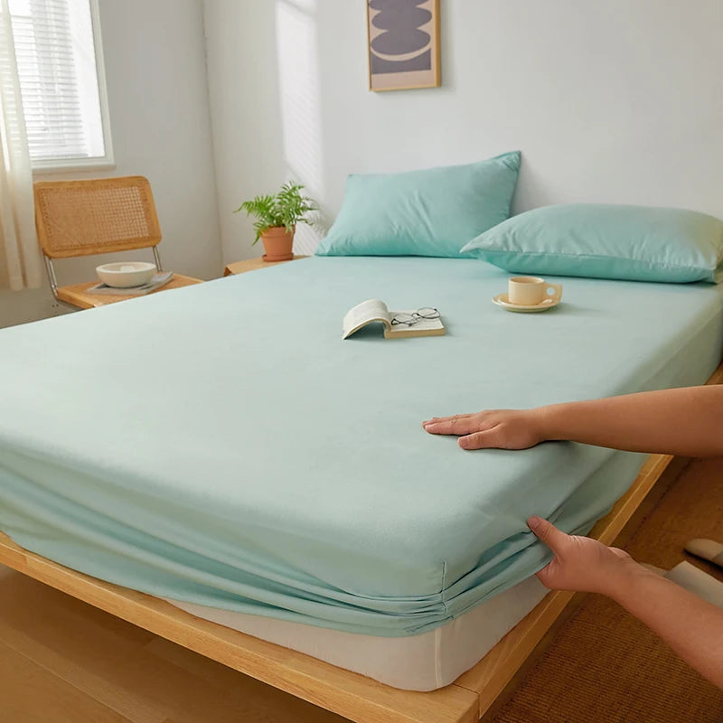 A light mint green fitted sheet is laid out on a wooden bed frame with a hand touching the fabric.
