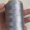 A spool of silver gray high tenacity polyester sewing thread is held in a hand, with a yellow plastic base visible.