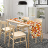 A light wood dining table set with a floral autumn leaf table runner featuring orange and white pumpkins.