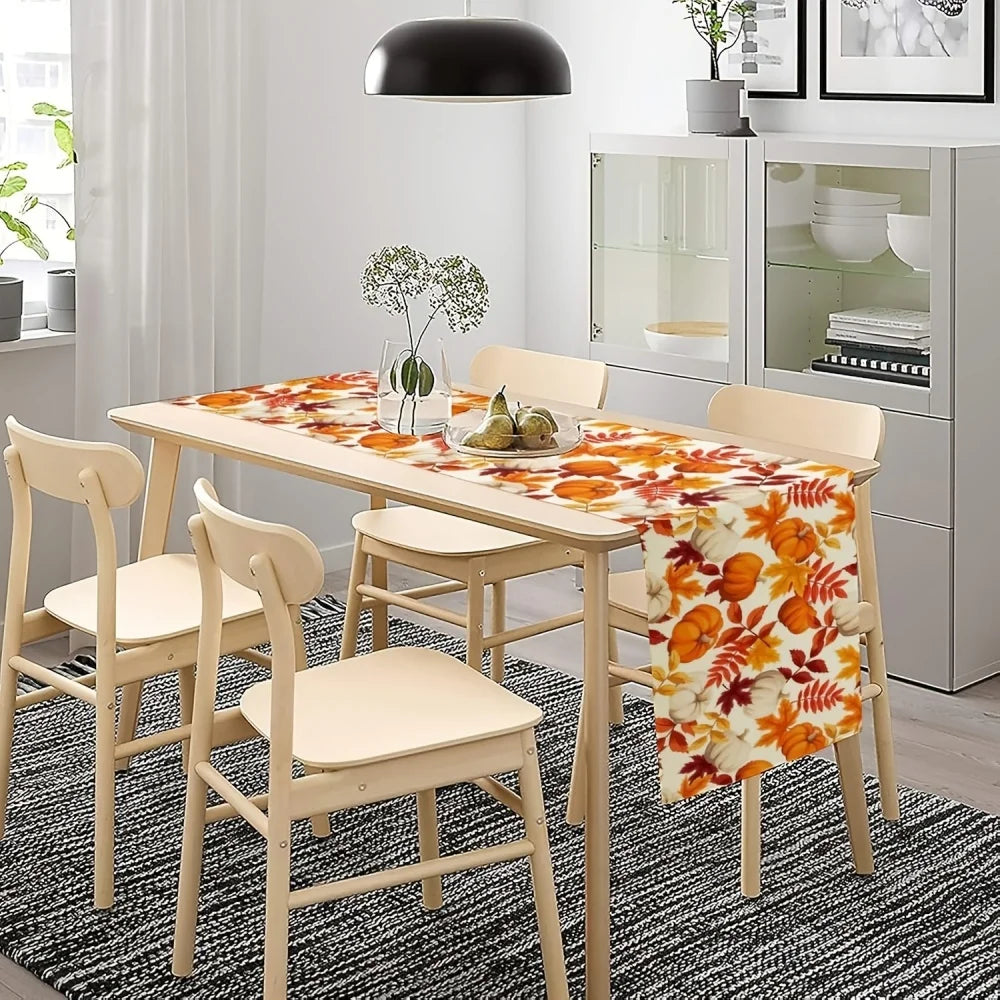 A light wood dining table set with a floral autumn leaf table runner featuring orange and white pumpkins.