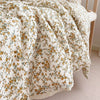 A close-up of a cream and gold floral print muslin blanket draped on a wooden surface.