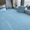 A light blue plush interlocking puzzle rug mat is displayed on a floor next to a window with a city view.