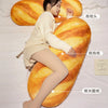 A woman is relaxing on a large, golden-brown plush baguette pillow in a bedroom setting.