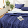 A navy blue quilted blanket is draped over a white bed with light blue bedding and green plants in the background.