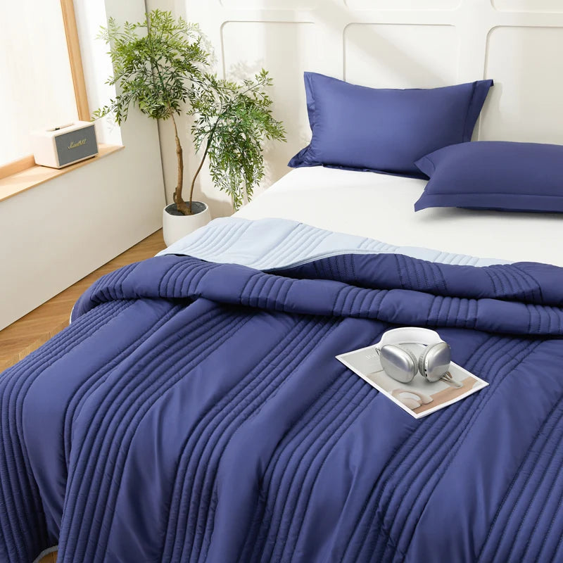 A navy blue quilted blanket is draped over a white bed with light blue bedding and green plants in the background.