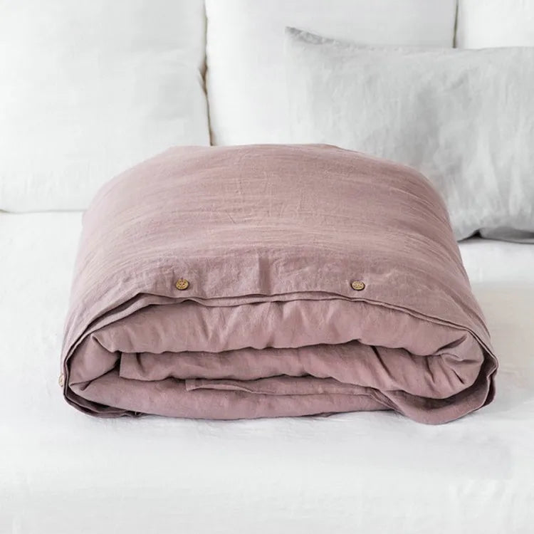 A folded mauve luxury French linen duvet cover resting on a white bed with white pillows.