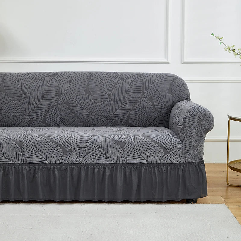 A charcoal gray jacquard stretch sofa cover featuring a leaf pattern displayed on a white floor.