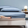 A hand is adjusting a smooth, dark blue bamboo bedding set on a white bed.