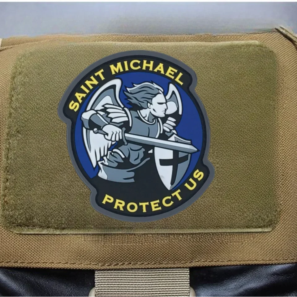 An embroidered patch featuring a winged figure and text that reads Saint Michael Protect Us on olive drab fabric.