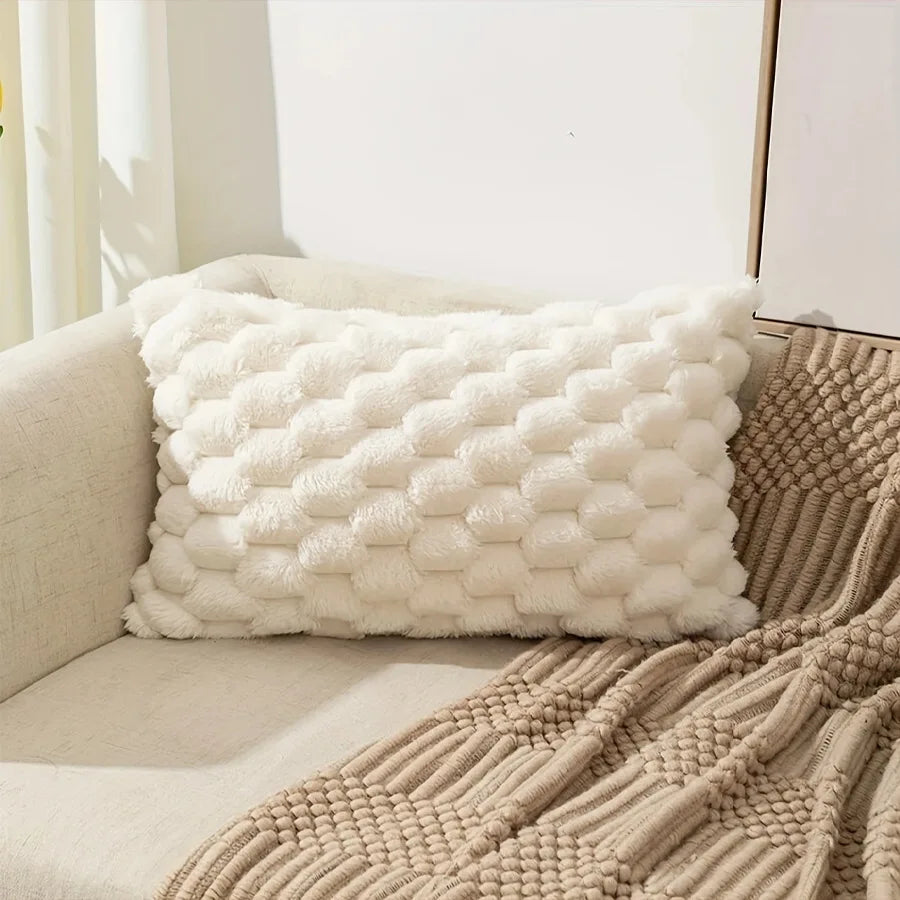 A fluffy white decorative plush pillow cover resting on a beige sofa next to a knitted throw blanket.