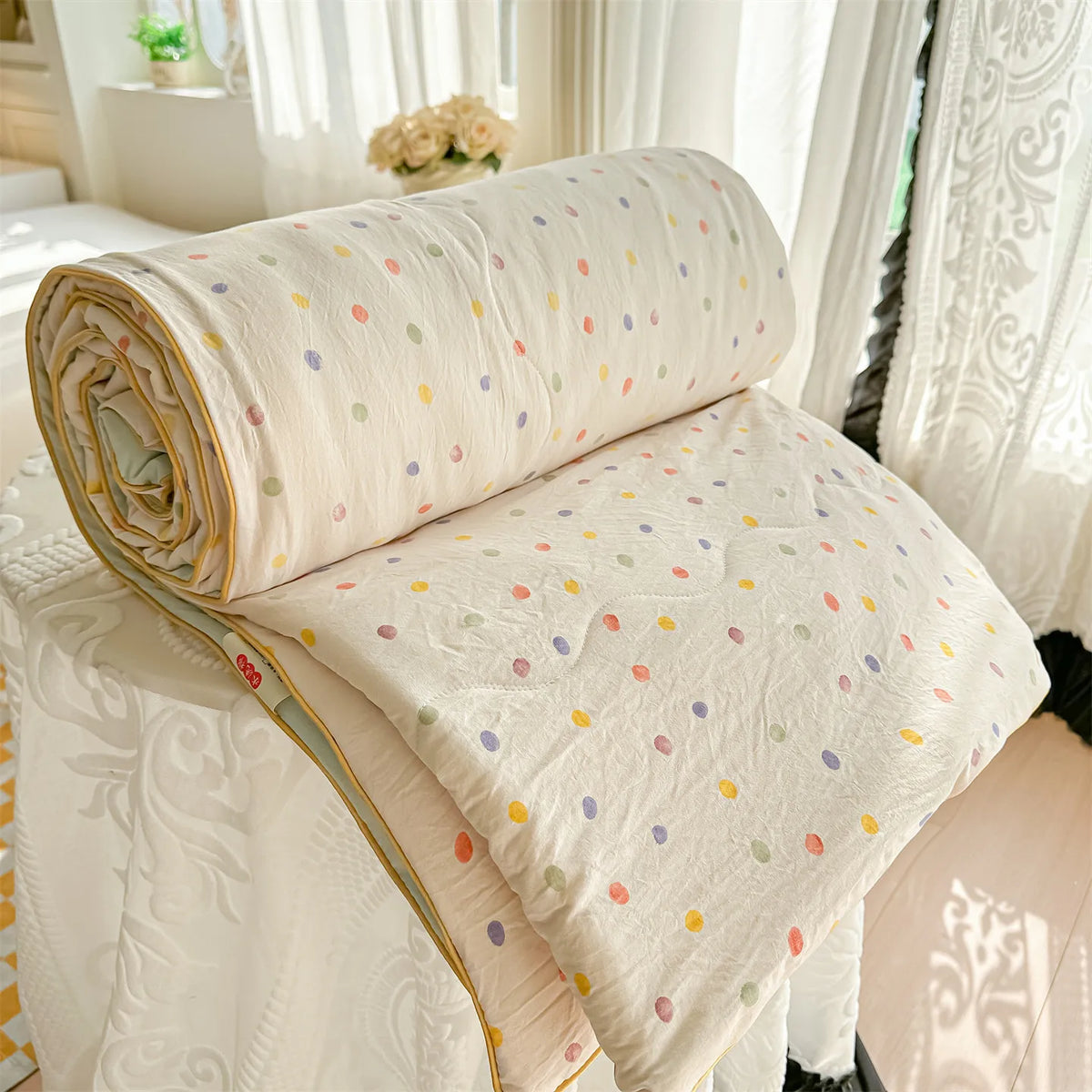 A rolled beige quilt blanket with multicolored polka dots resting on a white fabric surface near sheer curtains.