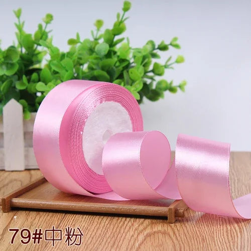 Pink satin ribbon roll with a pink ribbon unwound on a wooden surface surrounded by green foliage.