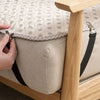 A hand is adjusting the elastic straps on a beige sheet corner gripper attached to a wooden bed frame.