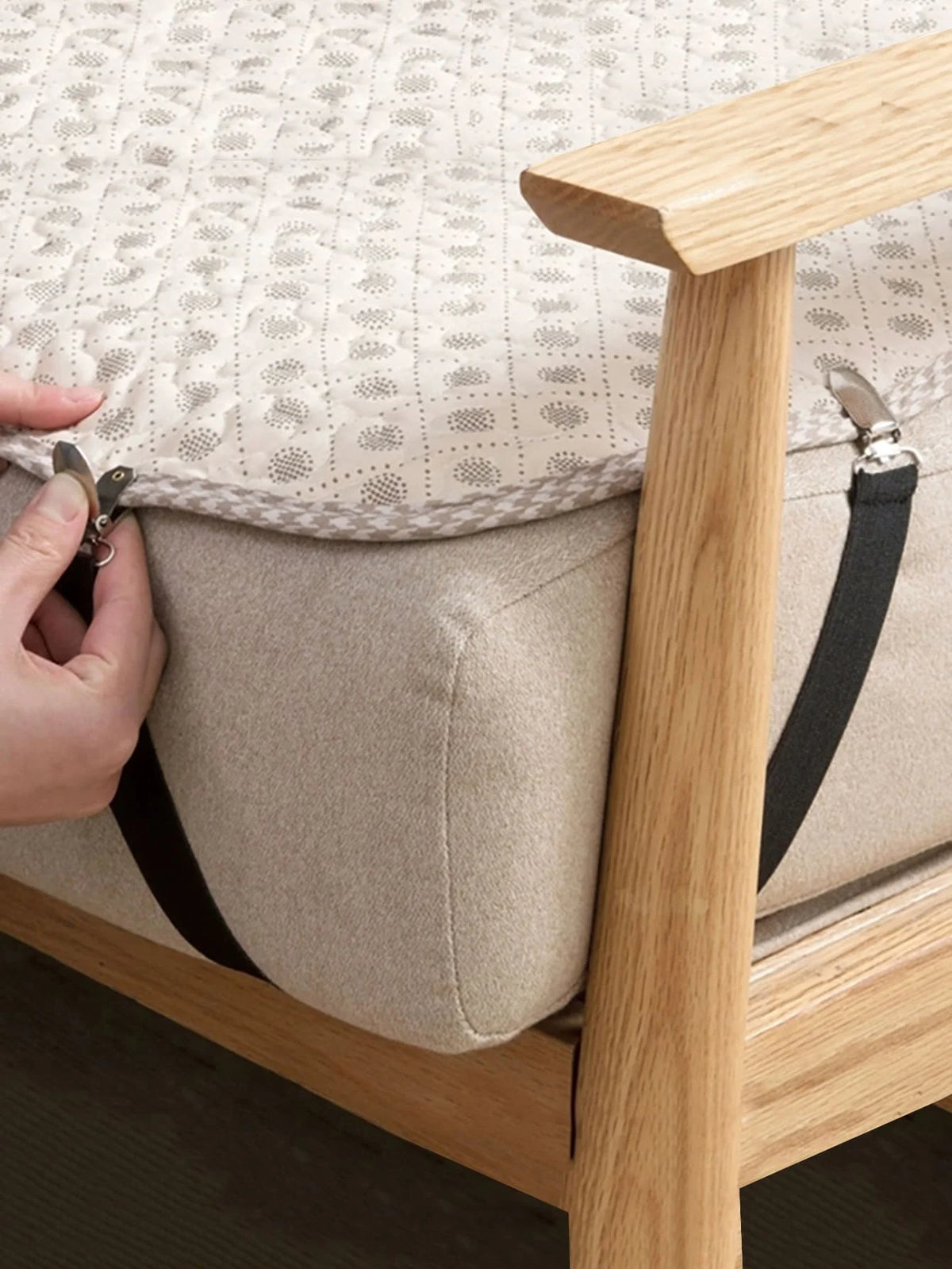 A hand is adjusting the elastic straps on a beige sheet corner gripper attached to a wooden bed frame.