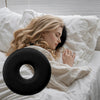 A blonde woman rests her head on a black comforting ear piercing pillow while lying in white bedding.