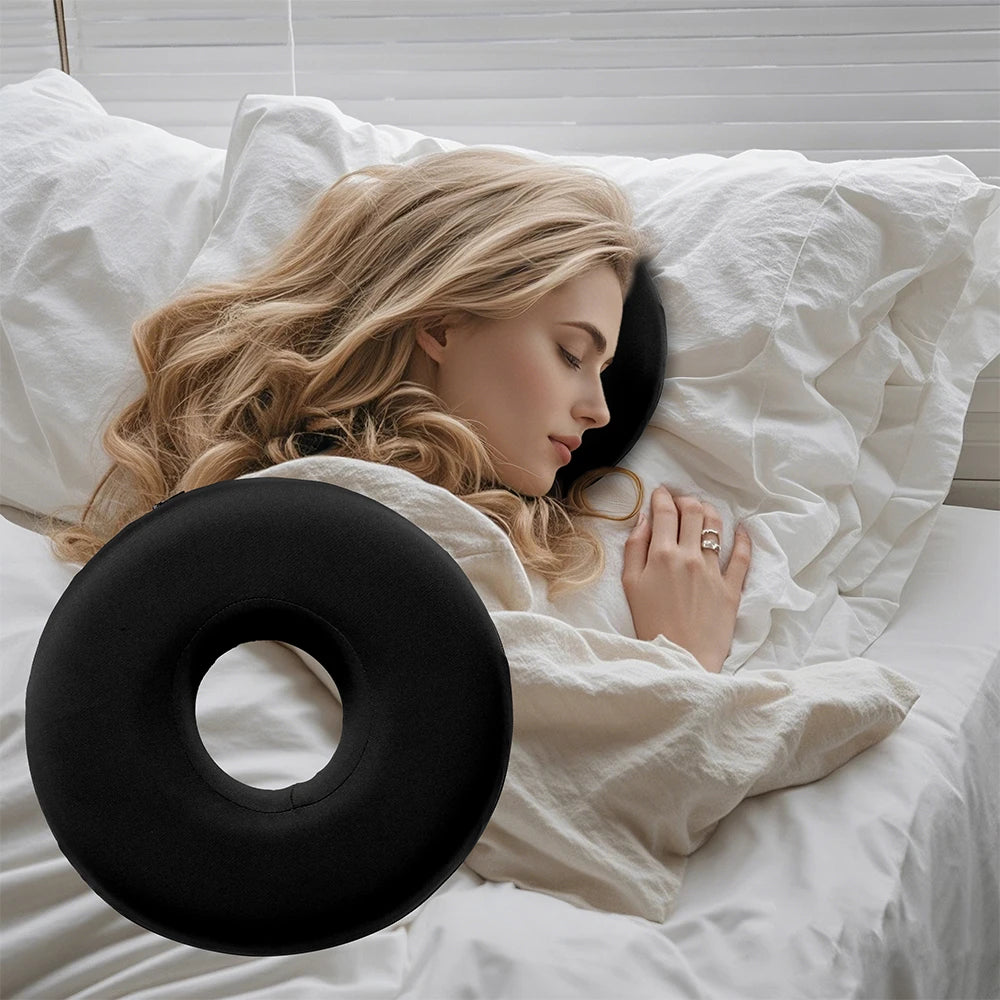 A blonde woman rests her head on a black comforting ear piercing pillow while lying in white bedding.