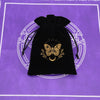 A black velvet pouch with gold foil butterfly and laurel design rests on a purple patterned surface.